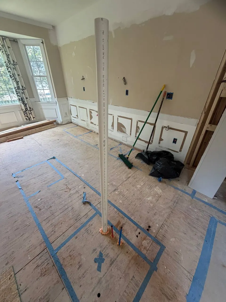 White PVC drain riser standing on the subfloor at the taped-out kitchen island location, with supply stub-outs at its base