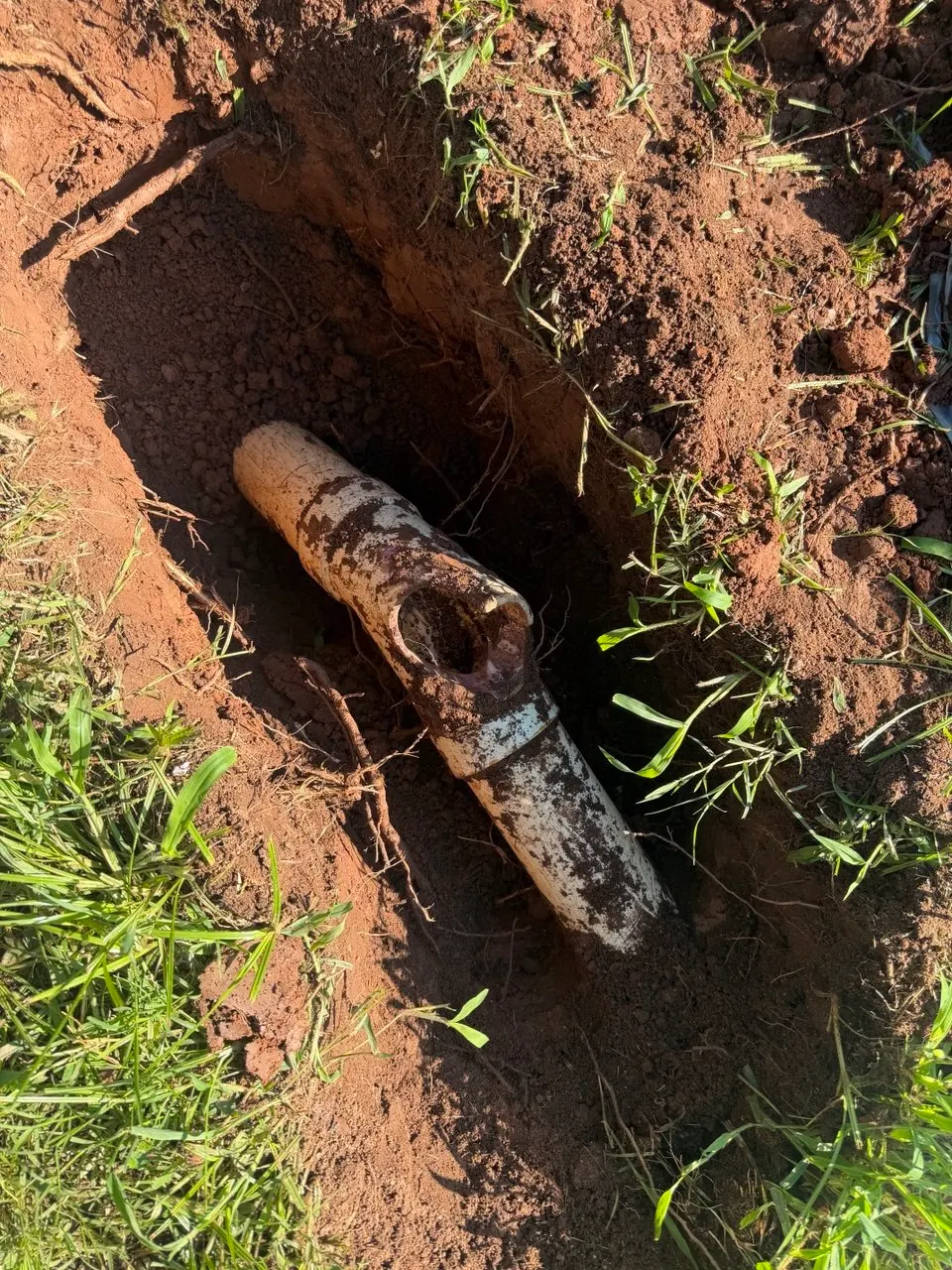 Broken white PVC sewer cleanout fitting exposed in an excavated hole in red clay soil