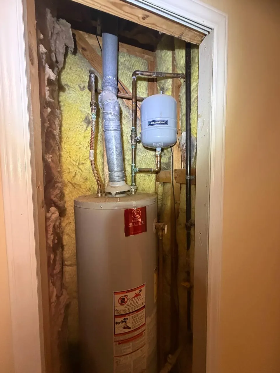 The previous gas water heater in the same closet before removal, with a corroded supply connector and the old expansion tank